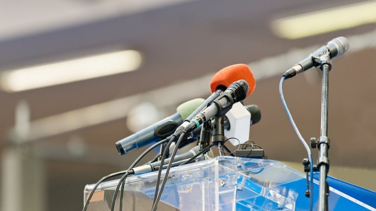 Multiple microphones in various colors are set up on a clear podium, ready for a speech or press conference. The background is blurred, focusing attention on the microphones. Multiple microphones in various colors are set up on a clear podium, ready for a speech or press conference. The background is blurred, focusing attention on the microphones.